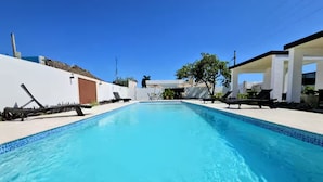 Piscina azul cristalina com espreguiçadeiras e um edifício branco ao fundo.