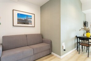 Ein Wohnzimmer mit einer Couch, einem gerahmten Bild an der Wand, einem Esstisch mit Stühlen und einer Vase mit Blumen.