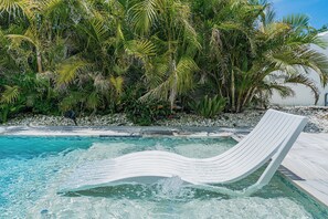 Ein Pool mit klarem Wasser, umgeben von üppigen tropischen Pflanzen und einer weißen Liege.