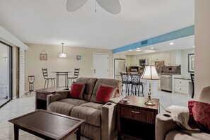 A living room with a sofa, coffee table, and lamp. There is a dining area with chairs and a table. The kitchen is visible in the background with appliances and cabinets. - 21 Sea Club Condo Near Beach (Indian Shores)