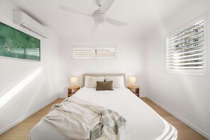Ein Schlafzimmer mit einem Bett, zwei Nachttischlampen, einem Deckenventilator, einem Fenster mit Jalousien und einem Bild an der Wand.