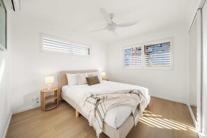 A bedroom with a bed, bedside lamps, a ceiling fan, and windows with blinds. - Ocean Glimpse Retreat - Sunlit Days by Kings Beach (Caloundra Region)