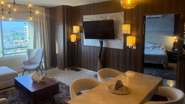 A modern hotel room with a large bed, a TV, a dining area, and a view of the city. - Luxury Apartment 40 m from the Beach and Bike Path (Mazatlán)