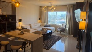 A modern living room with a sofa set, a dining area with chairs, a TV, and a large window with curtains. - Luxury Apartment 40 m from the Beach and Bike Path (Mazatlán)