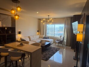 A modern living room with a sofa set, a dining area with chairs, a TV, and a large window with curtains. - Luxury Apartment 40 m from the Beach and Bike Path (Mazatlán)