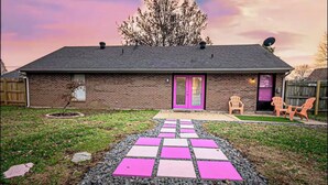 Une maison en briques avec une porte rose, une antenne parabolique et un chemin pavé menant à un espace extérieur aménagé pour s’asseoir.