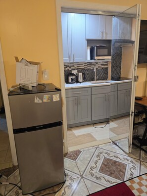 A compact kitchen with grey cabinets, a stainless steel refrigerator, a microwave, and a tiled backsplash.