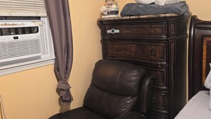Une pièce comprenant un fauteuil en cuir marron, une commode en bois, un lit et une fenêtre avec des stores et un rideau.