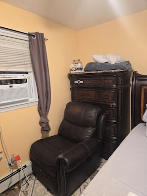 A room with a brown leather chair, a wooden dresser, a bed, and a window with blinds and a curtain.