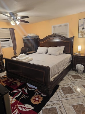 A bedroom with a wooden bed, a ceiling fan, a nightstand, a lamp, and a window with blinds.