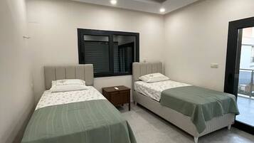 Un dormitorio con dos camas, una ventana con persianas, una lámpara en el techo y una mesita de noche pequeña.
