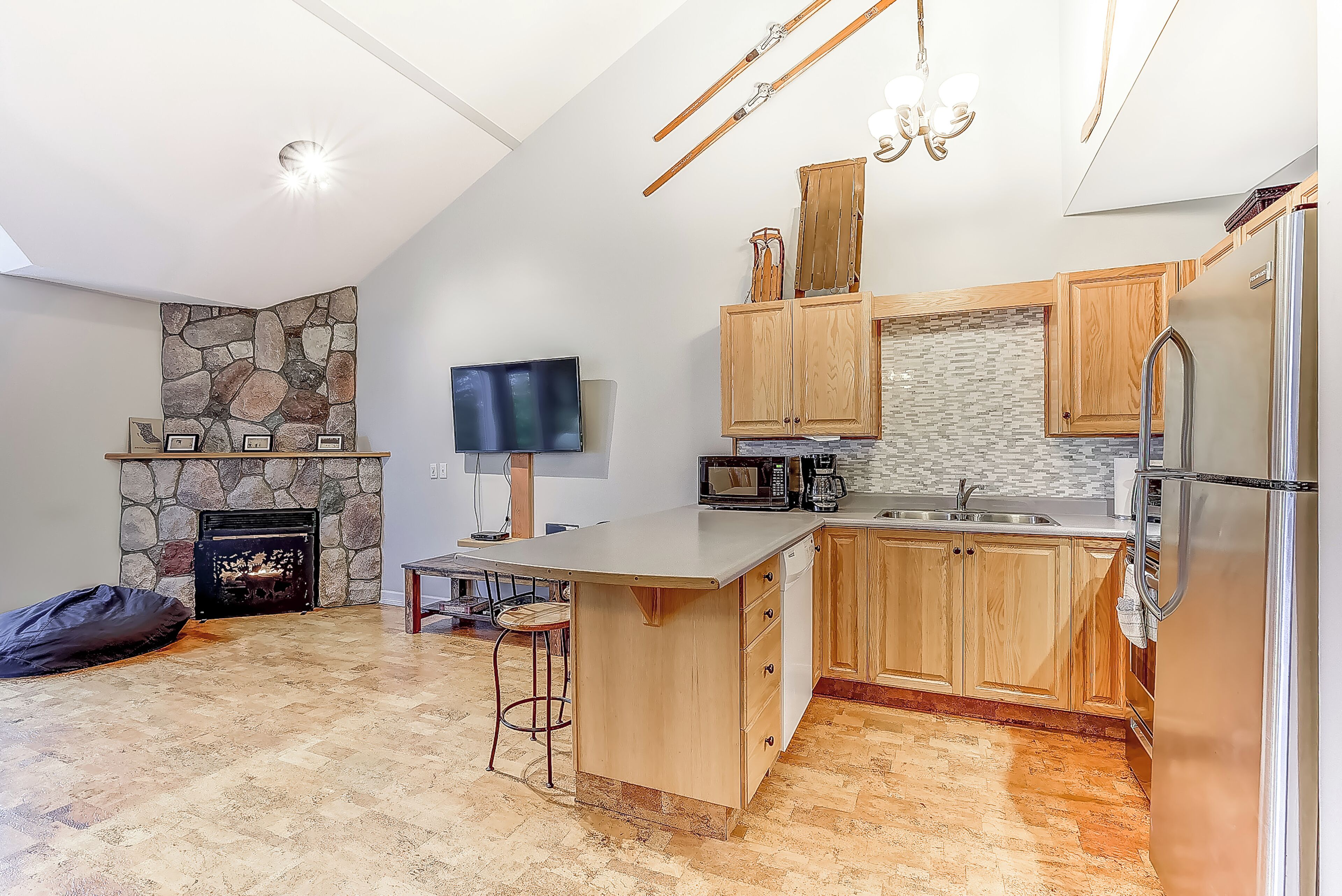 A modern kitchen with a stone fireplace, wooden cabinets, a stainless steel refrigerator, and a bar area.