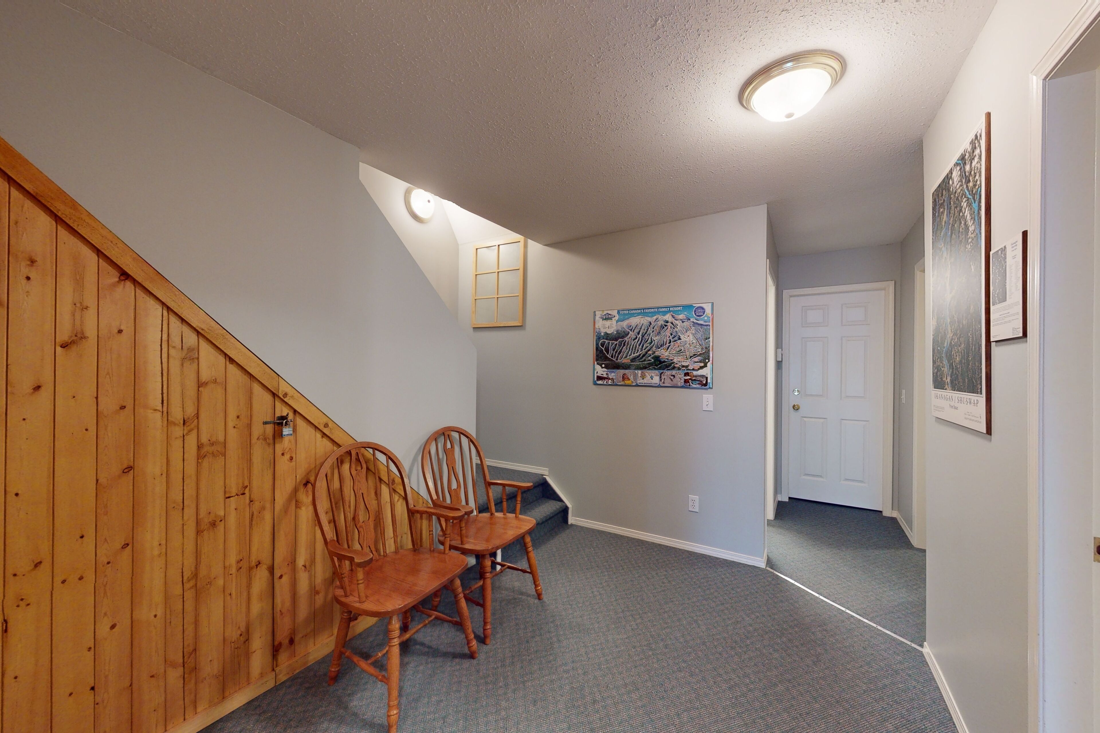 A hallway with a wooden staircase, two wooden chairs, a framed map, and a door.