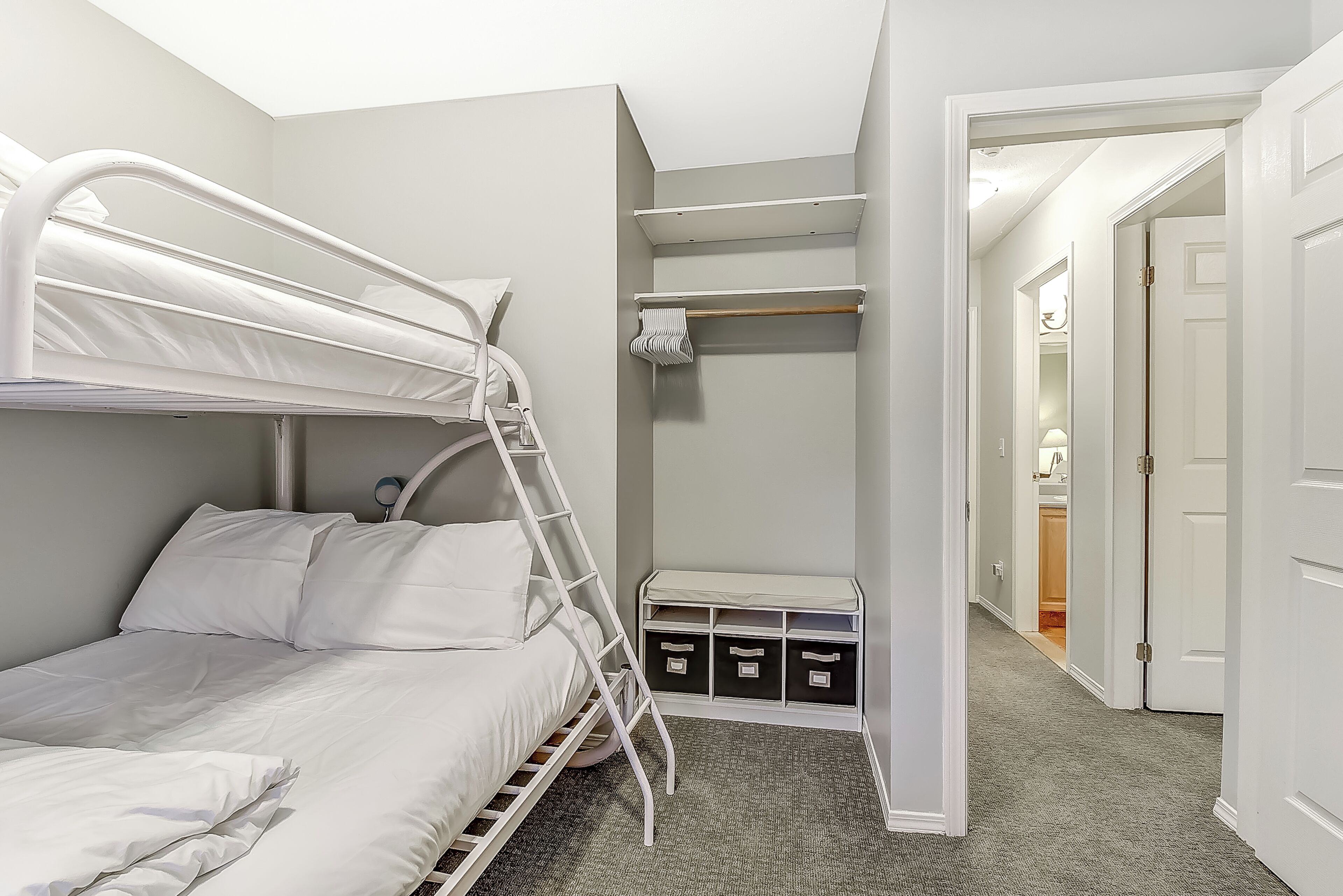 Un lit superposé avec une échelle, un petit meuble de rangement avec des tiroirs et un couloir menant à une porte.