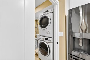 A laundry room with stacked front-loading washers and dryers, a water heater, and a stainless steel refrigerator. - Aransas Princess #314, Remodeled Gulf-Front 2BR + Den, Sleeps 6 (Port Aransas)