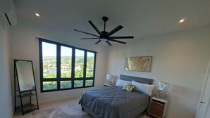 Ein Schlafzimmer mit großem Fenster, Deckenventilator, Bett mit grauer Bettdecke, Nachttische mit Lampen und einem an der Wand hängenden Kunstwerk.
