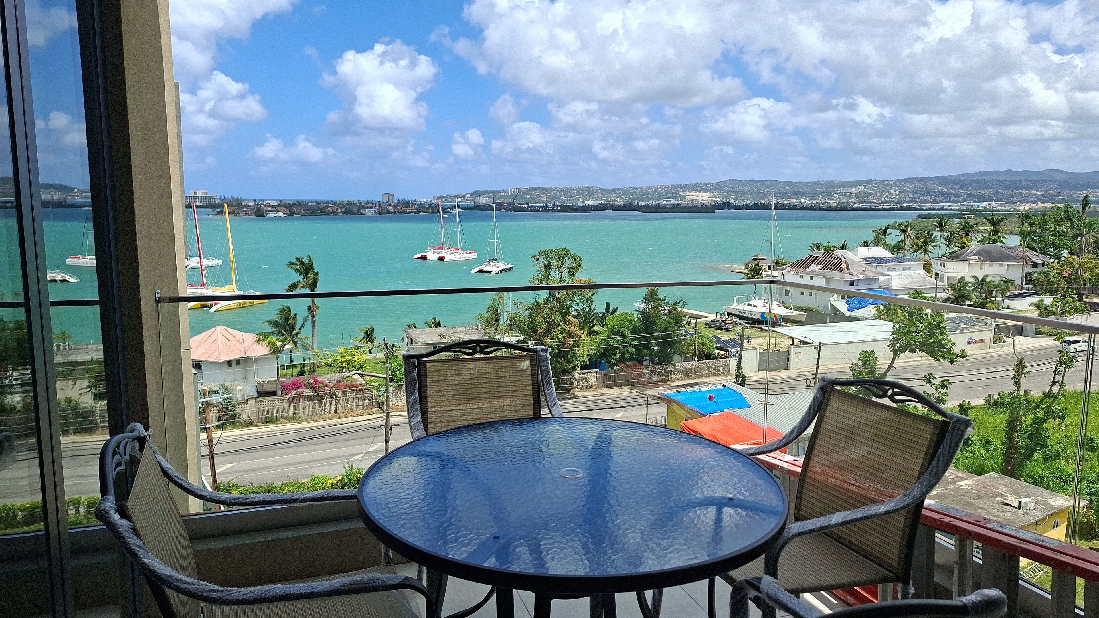 En balkon med et glasbord og stole med udsigt over en lysthavn med både og bybillede.