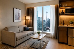 Une chambre d’hôtel moderne avec un canapé, une petite table, une vue sur le paysage urbain, un coin cuisine et un tableau encadré au mur.