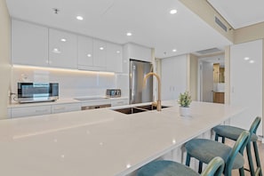 Eine moderne Küche mit weißen Schränken, einem Edelstahl-Kühlschrank, einer Mikrowelle, einem Toaster und einer Spüle mit goldenem Wasserhahn. An der Kochinsel stehen zwei blaue Polsterstühle.