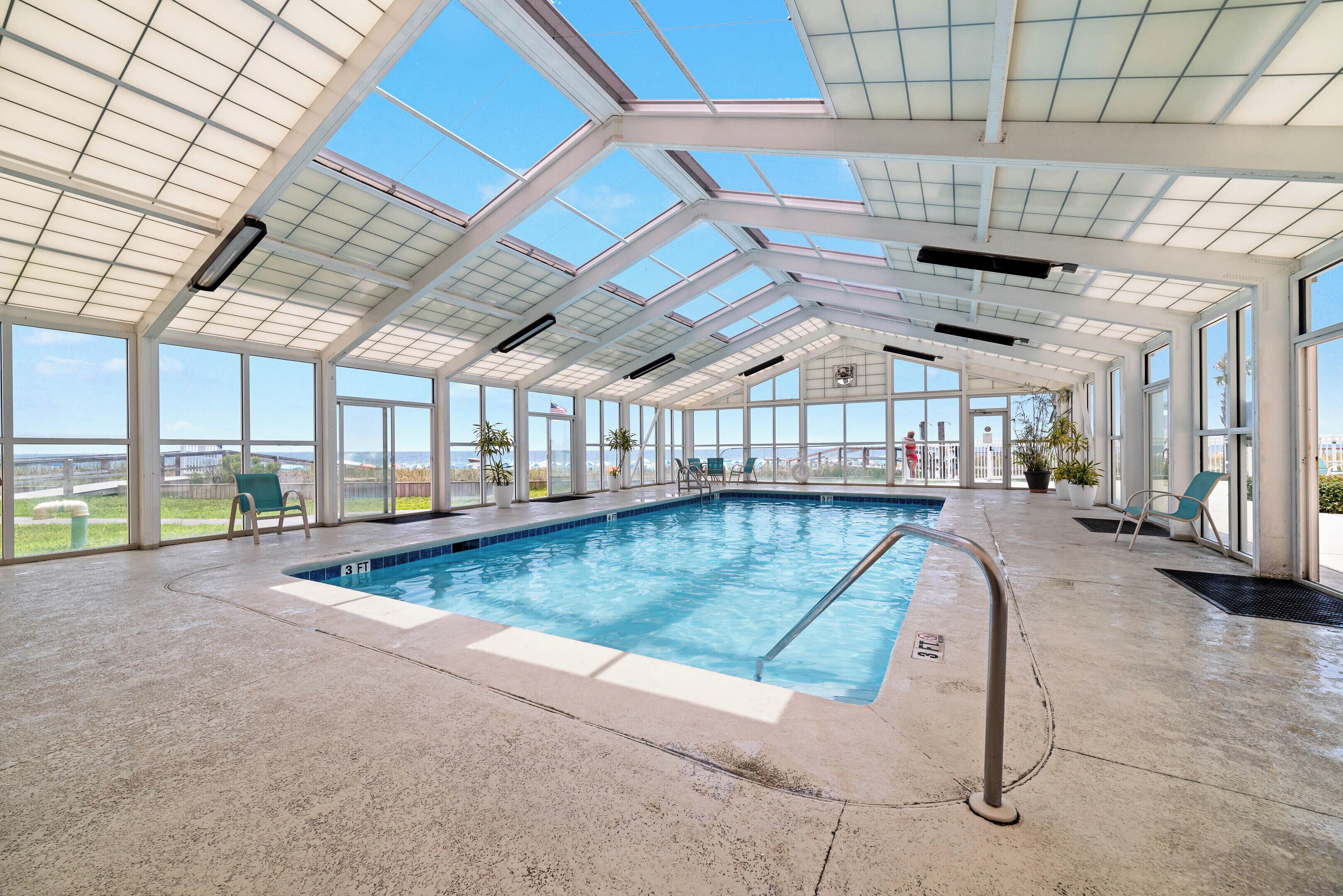 An indoor swimming pool with a glass ceiling, clear views of the outside, and a few lounge chairs.