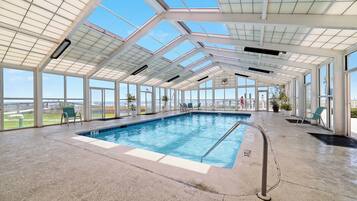 An indoor swimming pool with a glass ceiling, clear views of the outside, and a few lounge chairs.