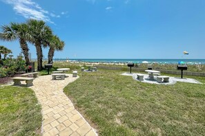 A paved pathway leading to a seating area with benches, surrounded by grass and palm trees, overlooking a beach with umbrellas and a clear sky. - Perdido Sun 216 | 4 Bdrm Corner Condo | Beachfront | Pools | Walk Everywhere | My Beach Getaways (Pensacola)