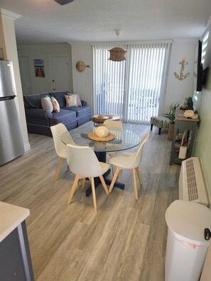 Ein modernes Wohnzimmer mit einem Glastisch, weißen Stühlen, einer Couch und einer Küchenzeile inklusive Kühlschrank.