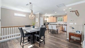 A spacious dining area with a chandelier, a kitchen in the background, and a wooden floor. - Blue Heron on May Vacation Rental (St. Augustine)
