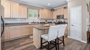 A kitchen with beige cabinets, a central island with two white bar stools, a microwave, and a stove. - Blue Heron on May Vacation Rental (St. Augustine)