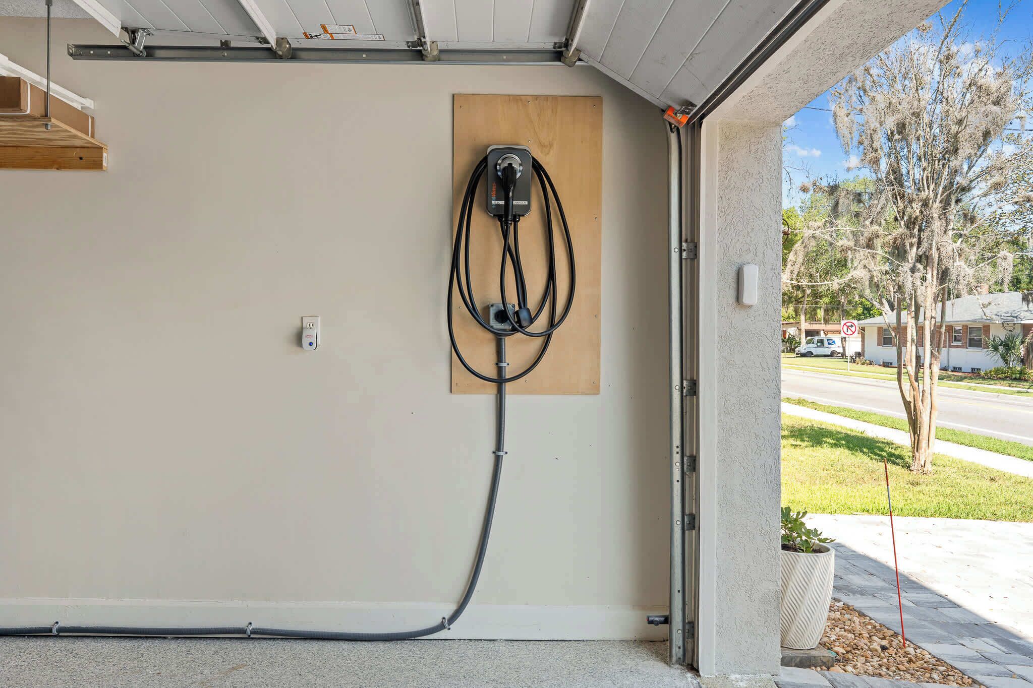 Un garage équipé d’une borne de recharge fixée au mur, d’une étagère en bois et d’une plante en pot à l’extérieur.