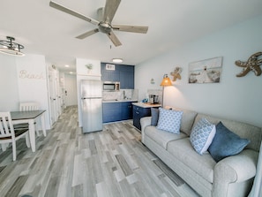 Eine moderne Küche mit Kühlschrank, Mikrowelle und Herd, ein Essbereich mit Tisch und Stühlen, ein Wohnzimmer mit Sofa und Kissen sowie ein Deckenventilator.