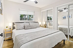 Ein Schlafzimmer mit einem großen Bett, Nachttischen mit Lampen, einem Deckenventilator, einem Fenster mit Blick auf Bäume und einer Glastür, die in einen anderen Raum führt.