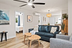 Ein modernes Wohnzimmer mit einem dunkelbraunen Ledersofa, zwei Holztischen, einem grauen Sessel und einem Deckenventilator.