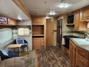 Een compacte camperkeuken met houten kastjes, een koelkast, een spoelbak en een eethoek met een stoel en tafel.