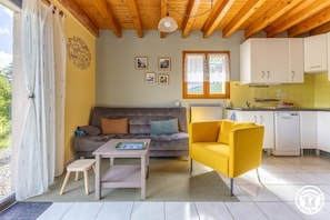 Ein gemütliches Wohnzimmer mit einer Couch, einem gelben Sessel, einem kleinen Tisch und einer Küchenzeile mit weißen Schränken und einem Geschirrspüler.