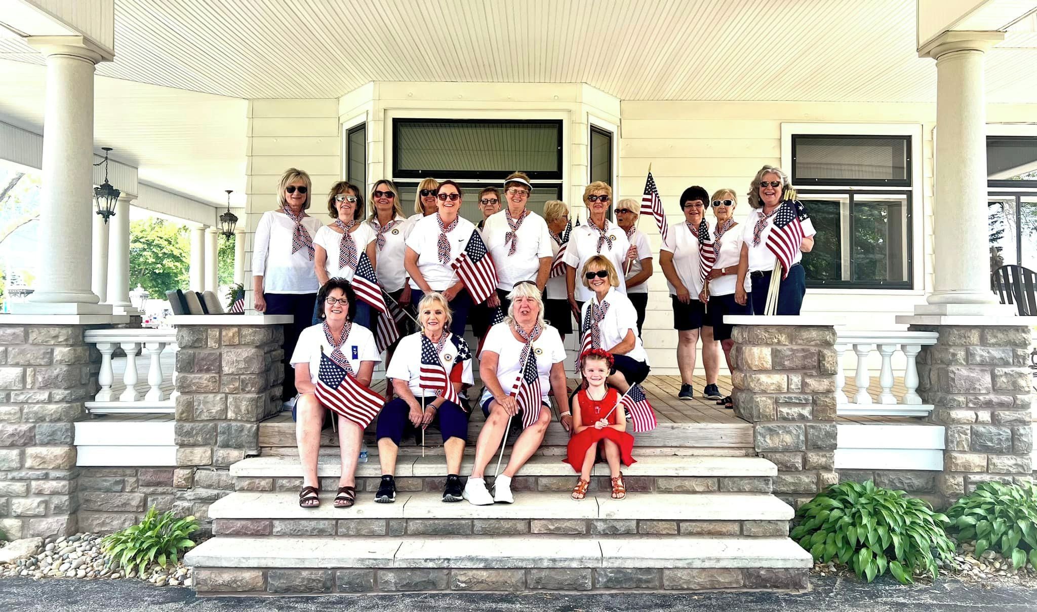 A group of people on a porch with stone steps and a covered area, some holding American flags.