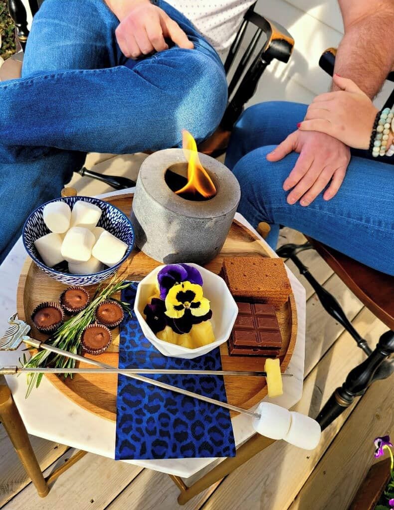 A tray with various food items, including marshmallows, chocolate, and flowers, on a wooden table outdoors.