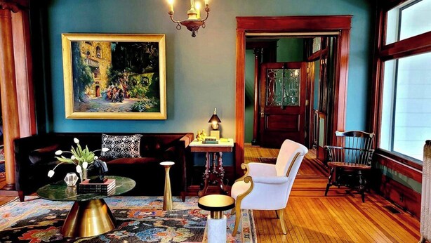 A room with a large painting, a chandelier, a sofa, a chair, a table, and a patterned rug. - 85Ten Hotel and Events  (Port Austin)