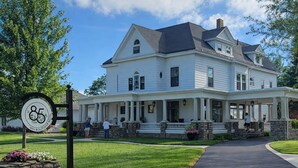 A large, two-story white building with a porch, stone steps, and a sign indicating it is a boutique hotel. - 85Ten Hotel and Events  (Port Austin)