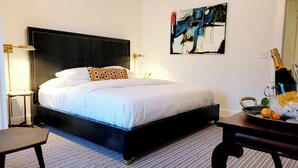 A hotel room with a large bed, a bedside table, a chair, a painting on the wall, and a bottle of champagne on a tray. - 85Ten Hotel and Events  (Port Austin)