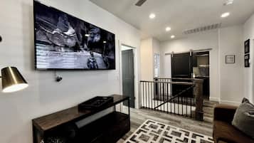A modern living room with a mounted TV, a dark wooden entertainment unit, a patterned rug, a brown sofa, and a staircase leading to a kitchen area.