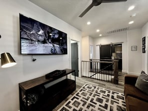 A modern living room with a mounted TV, a dark wooden entertainment unit, a patterned rug, a brown sofa, and a staircase leading to a kitchen area.