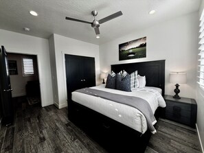 A bedroom with a bed, a ceiling fan, a nightstand with a lamp, a painting on the wall, and a door with a sign.