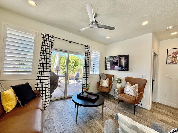 A living room with a sofa, chairs, a coffee table, a ceiling fan, and a TV mounted on the wall.