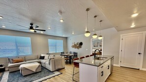 A modern living space with a sofa, dining table, kitchen island, ceiling fan, recessed lighting, and a door.
