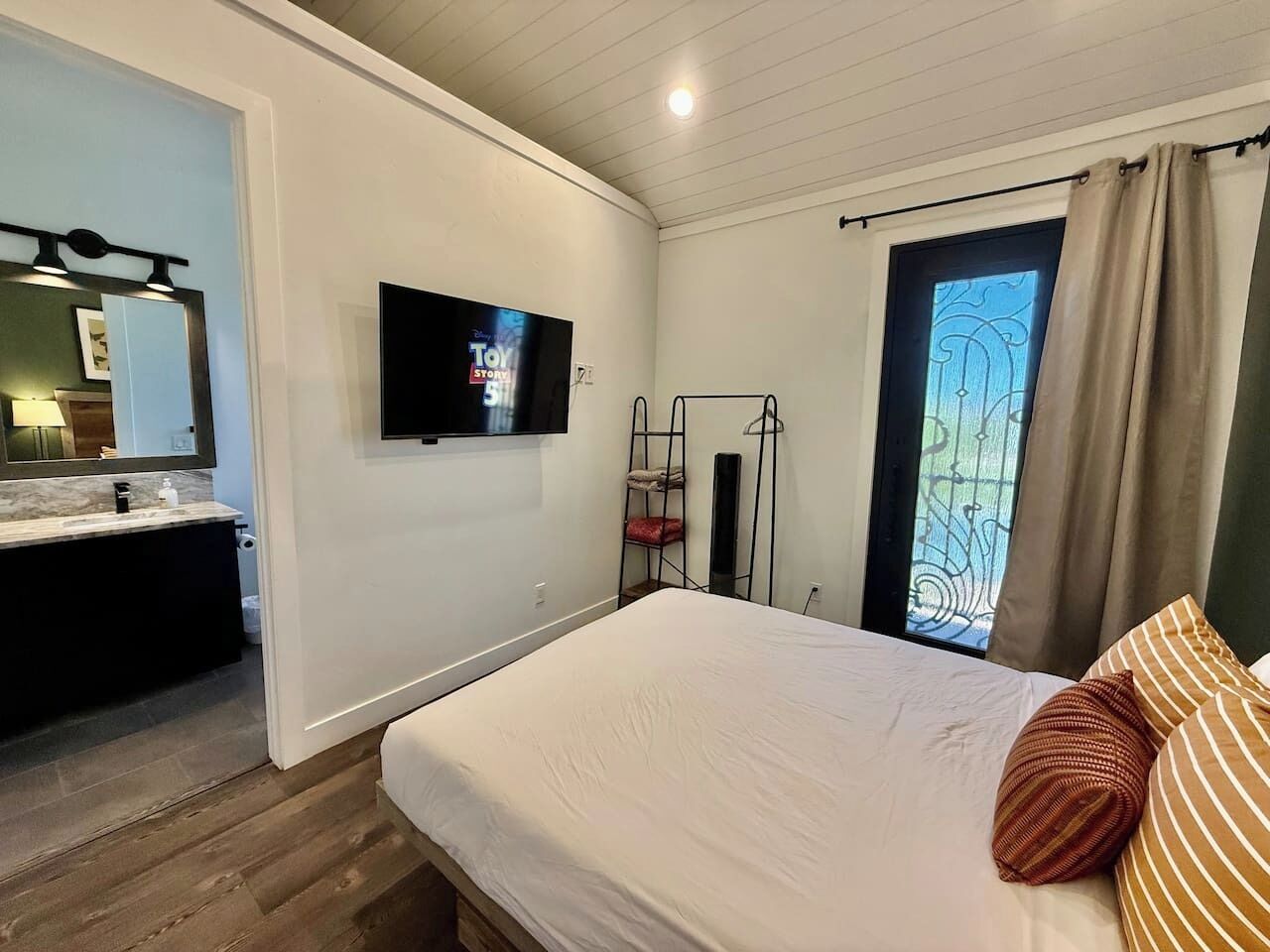 A bedroom with a bed, a TV, a window, and a bathroom with a sink and mirror.