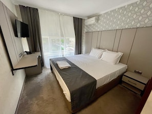 Una habitación de hotel con una cama, una mesita de noche, un televisor montado en la pared y una ventana con cortinas.