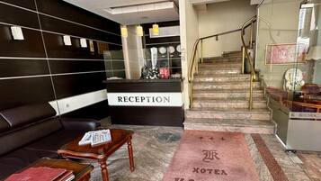 Zona de recepción de un hotel con mostrador, un sofá, una mesa de centro y una escalera que conduce al piso superior.