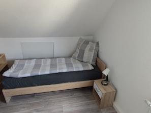 A small, single-bed room with a wooden headboard, a bedside table with a lamp, and a nightstand with a power outlet.