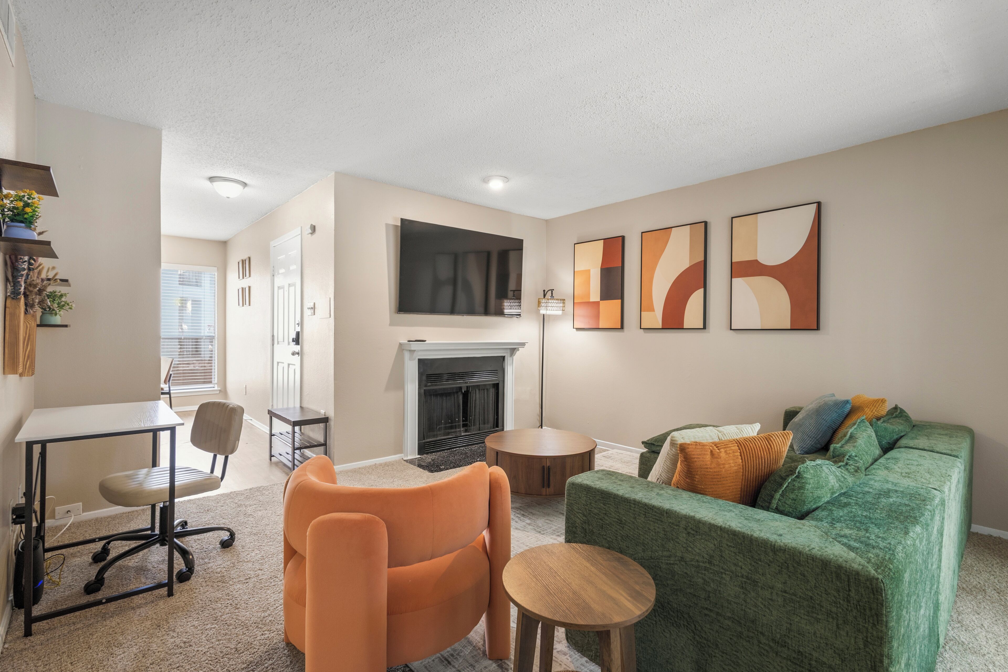A living room with a fireplace, a green sofa, an orange chair, a small table, and abstract wall art.